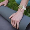 A person's hands are shown rolling up a yoga mat outdoors, with grass in the background. The person is wearing a gold and cream smartwatch. Náhled obrázku produktu