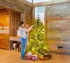 A woman is holding a child and decorating a Christmas tree in a room with wooden walls. Produktbild-Vorschau