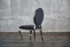 A black velvet chair with a silver frame stands against a textured gray wall and a dark wooden floor. Produktbild-Vorschau