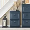 A dark blue shoe cabinet with two drawers is shown next to a staircase, with a lantern and decorative items on top. Náhľad obrázku produktu