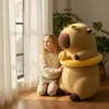 A young girl kneeling next to a large capybara plush toy in a room with natural light coming from a window. Anteprima dell’immagine prodotto
