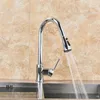 A chrome kitchen faucet with a pull-out spray head is shown in operation, with water flowing into a stainless steel sink. The background consists of beige tiled walls. Náhľad obrázku produktu