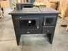 A black wood-burning stove with a metal plate on top, shown in a warehouse or workshop setting. Anteprima dell’immagine prodotto