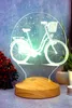 A 3D LED lamp shaped like a bicycle is displayed on a wooden base, with decorative elements in the background. Produktbild-Vorschau