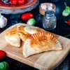 A close-up shot of several golden-brown empanadas on a wooden cutting board, with some ingredients like tomatoes and garlic visible in the background. Produktbild-Vorschau