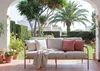 An outdoor sofa on a tiled patio, surrounded by lush greenery and palm trees, with a white building in the background. Podgląd zdjęcia produktu