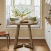 A round wooden dining table with a vase of flowers and decorative items on it, placed in front of a window seat with cushions. Anteprima dell’immagine prodotto