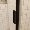 A close-up view of a black shower door handle against white tiled walls. Produktbild-Vorschau