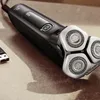 A close-up shot of a black electric shaver with silver shaving heads, placed on a textured surface. Náhled obrázku produktu