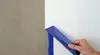 A hand is applying blue painter's tape to a wall where two different colors meet. Anteprima dell’immagine prodotto