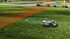 A robotic lawnmower is shown on a green lawn, with a swimming pool and a house in the background. Orange and blue highlights indicate the mower's operational area and path. Voir les images du produit