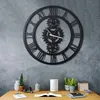 A large, black, gear-themed wall clock with Roman numerals hangs on a textured gray wall above a wooden desk. The desk has various decorative items and books. Náhľad obrázku produktu