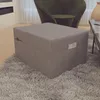 A gray ottoman is placed on a fluffy rug in a living room setting, with a green chair and a coffee table visible in the background. Produktbild-Vorschau