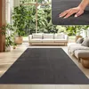 A living room with a sofa and a large rug in front of a window with a view of trees. Produktbild-Vorschau