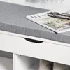 A close-up view of a white storage bench with a gray upholstered seat, featuring a magazine and glasses on top and a drawer opening. Anteprima dell’immagine prodotto