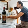 A man drinks from a glass in a kitchen, with a bottle of Amer Picon on the counter. Anteprima dell’immagine prodotto
