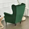 A green velvet wingback chair is shown from the back, with a woven stool and a plant in the background. Anteprima dell’immagine prodotto