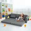 A young girl is laughing on a gray modular sofa in a playroom with shelves full of toys. Náhled obrázku produktu
