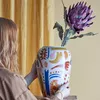 A person holding a decorative vase with a flower in it, with a yellow curtain in the background. Náhled obrázku produktu