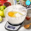 A digital kitchen scale with a bowl of flour and eggs on top, surrounded by baking ingredients and a recipe book. Produktbild-Vorschau
