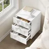 A white nightstand with three open drawers, placed next to a bed and near a window. Produktbild-Vorschau