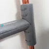 A close-up shot of gray pipe insulation on copper pipes, attached with clips, against a white wall. Produktbild-Vorschau
