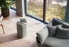 An air purifier is placed on a wooden floor next to a sofa in a modern living room with a large window overlooking a city view. Podgląd zdjęcia produktu