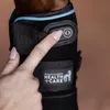 A close-up of a person's finger pressing a button on a black horse leg wrap with blue trim. The Waldhausen Health + Care logo is visible. Podgląd zdjęcia produktu