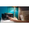 A person's hand is shown interacting with a black digital radio on a white nightstand, next to a bedside lamp and a bed. Produktbild-Vorschau