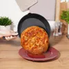 A hand is holding a cast iron skillet with a pizza inside, tilting it over a red plate on a wooden table. Náhled obrázku produktu