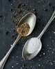 Two spoons are shown, one filled with salt and the other with ground pepper, against a dark background with scattered peppercorns and salt crystals. Produktbild-Vorschau