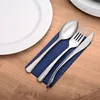 A set of cutlery is laid out on a blue napkin on a wooden table, next to a white plate. Produktbild-Vorschau