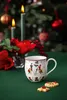 A Christmas-themed mug with a snowman design sits on a green tablecloth, with candles and festive decorations in the background. Produktbild-Vorschau