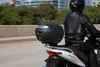 A person riding a scooter on a road with buildings and trees in the background. Anteprima dell’immagine prodotto