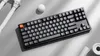 A black mechanical keyboard with gray keycaps is placed on a light gray surface, with some decorative white objects in the background. Produktbild-Vorschau