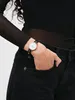 A person wearing a black top and black jeans, with a watch on their wrist. Produktbild-Vorschau
