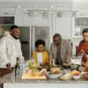 A family is gathered around a kitchen counter, preparing food. There are various dishes and ingredients laid out. Produktbild-Vorschau