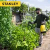 A man is using a yellow battery-powered sprayer in a garden with lush green plants. Podgląd zdjęcia produktu