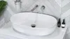 A white oval sink with chrome faucets and a chrome drain stopper, set against a herringbone tile backsplash in a modern bathroom. Podgląd zdjęcia produktu