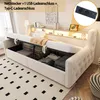 A beige upholstered sofa bed with storage, showing its open storage compartment filled with folded linens and blankets. The headboard has integrated lighting and shelving, and a close-up inset shows a power outlet with USB ports. Náhľad obrázku produktu
