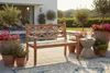 A wooden bench is placed on a patio next to a swimming pool, surrounded by potted plants and a side table with drinks. Produktbild-Vorschau