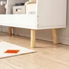 A close-up view of the bottom section of a white bookshelf with wooden legs, resting on a wooden floor. A rug with an orange square is partially visible in the foreground. Náhľad obrázku produktu