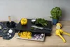 A black cutting board with a drawer containing sliced onions and yellow bell pepper strips. Ingredients like garlic, parsley, and lemons are arranged around it, along with a mortar and pestle and a lemon squeezer. Produktbild-Vorschau