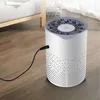 A white cylindrical air purifier with a gray decorative top is plugged in and sitting on a wooden floor next to a bed. Produktbild-Vorschau