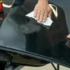 A person is using a cloth to wipe a black surface, possibly a car part, with a product bottle visible in the background. Anteprima dell’immagine prodotto