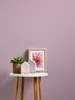 A small white table with decorative items on it, including a potted plant, two house-shaped objects, and a framed picture of a flower, against a plain purple background. Produktbild-Vorschau