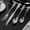 A set of silverware, including a fork, knife, and spoons, is displayed on a black surface. A white coffee cup and saucer are visible in the upper left corner. Produktbild-Vorschau