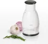 A white manual food chopper with a black lid sits on a white surface next to a head of garlic, a slice of red onion, and a sprig of rosemary. Produktbild-Vorschau