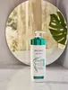A bottle of Biopoint body cream is placed in front of a circular mirror, reflecting a room with plants and curtains. Podgląd zdjęcia produktu