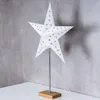 A white star-shaped lamp with gold star cutouts, standing on a wooden base, against a textured gray wall. Produktbild-Vorschau